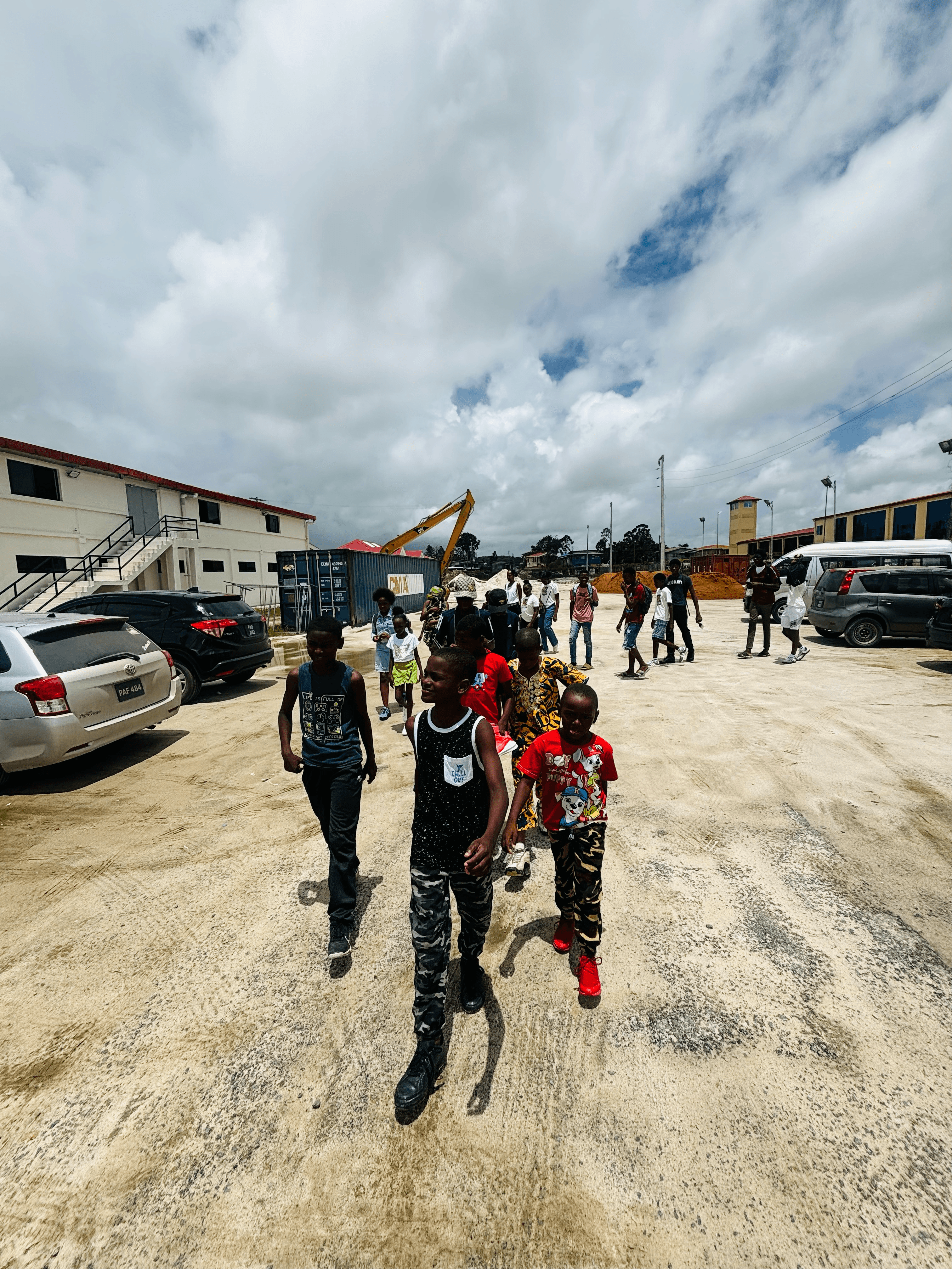 Children walk across a sandy lot past parked cars, a shipping container, and an excavator.